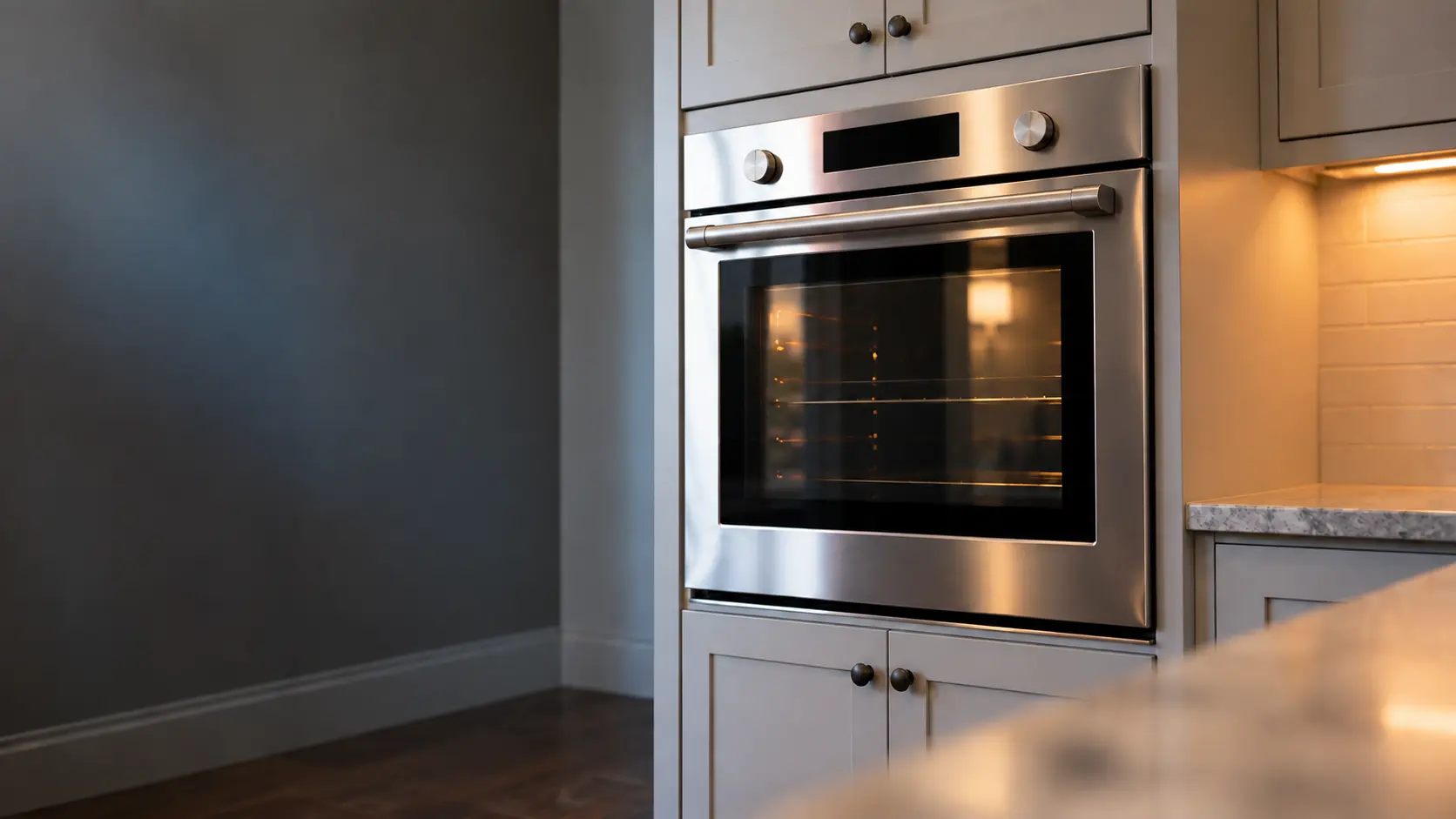 A kitchen wall with the oven in a two-flat kitchen with a wall oven in painted cabinets, seen from a low angle, the oven door glass catching the light.