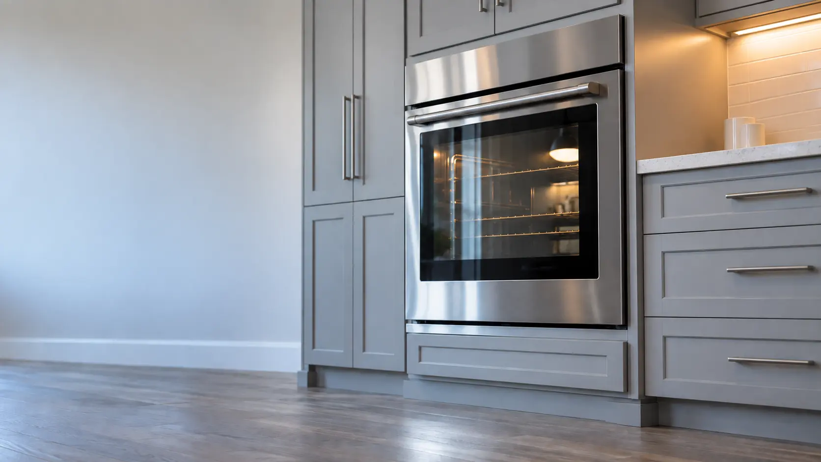 A kitchen wall with the oven in a newer subdivision kitchen with a wall oven in grey cabinets, seen from a low angle, the oven door glass catching the light.