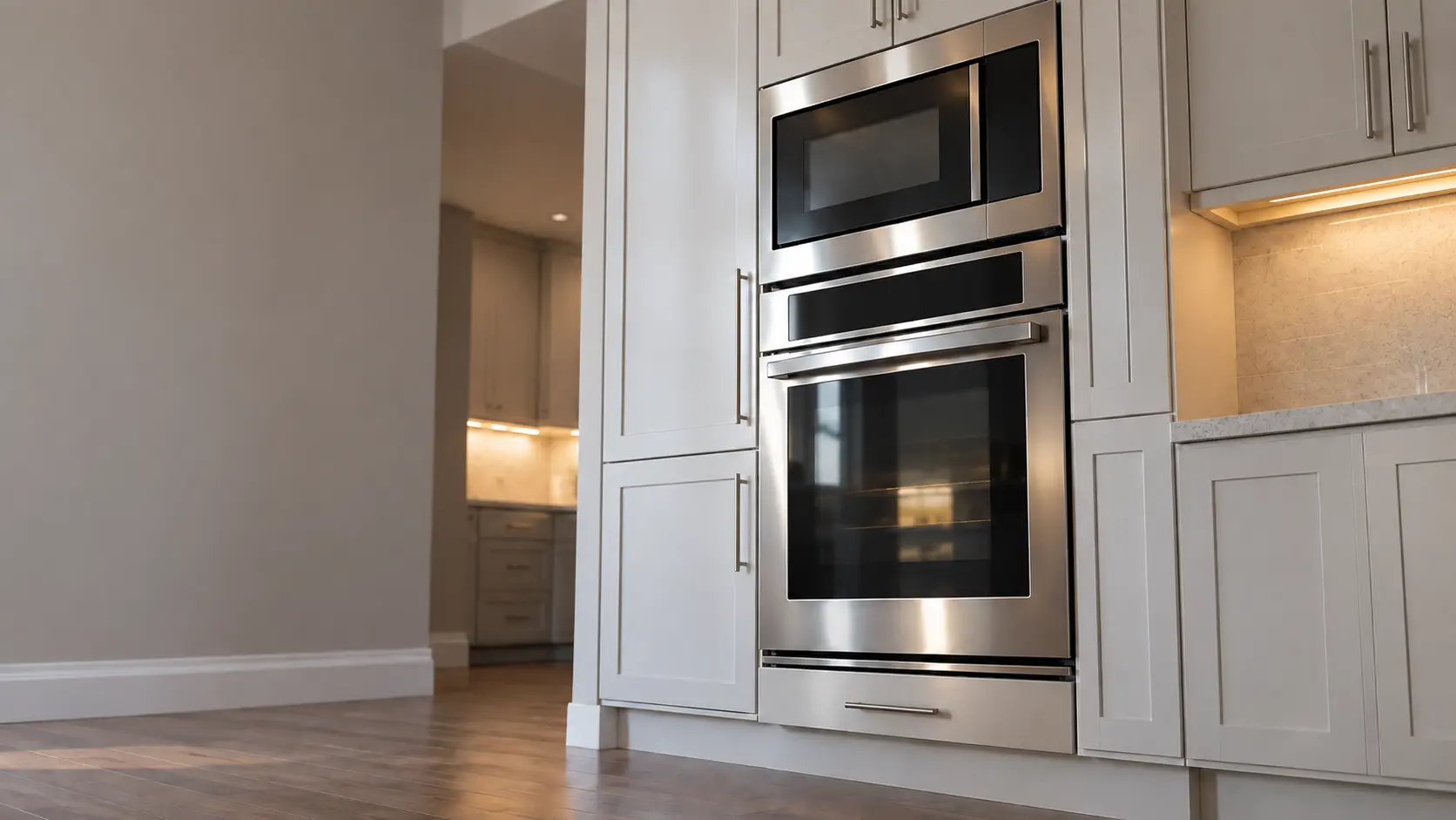 A kitchen wall with the oven in a master-planned community kitchen with a wall oven and microwave stack, seen from a low angle, the oven door glass catching the light.