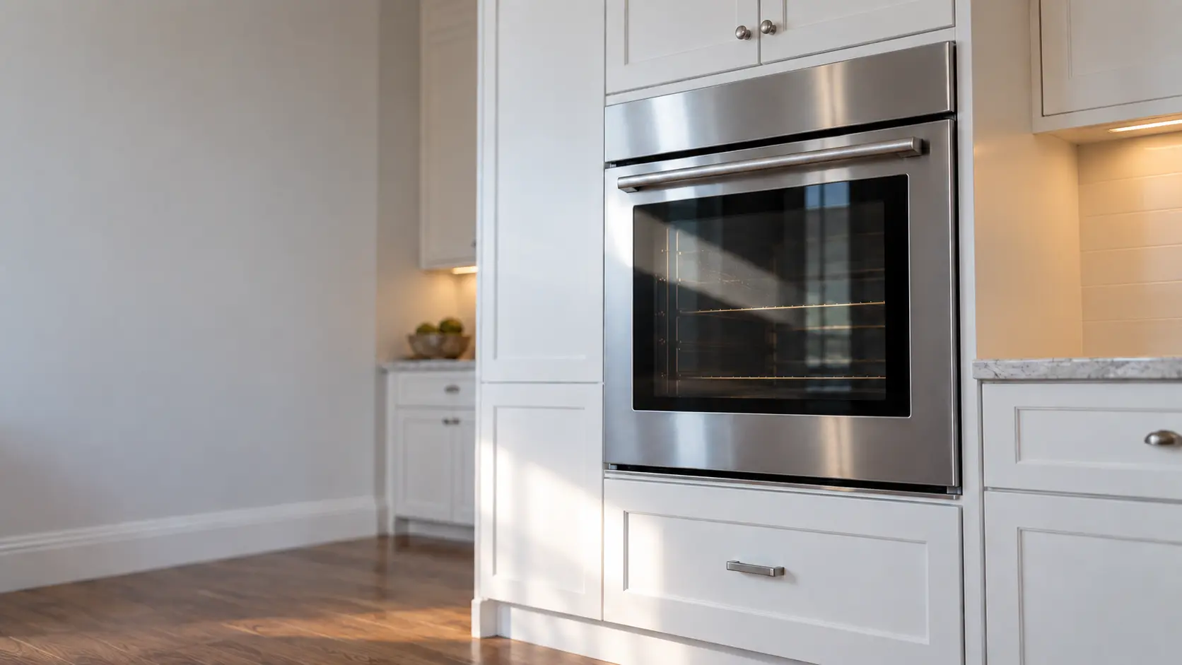 A kitchen wall with the oven in a shingled Long Island house kitchen with a wall oven in white cabinets, seen from a low angle, the oven door glass catching the light.