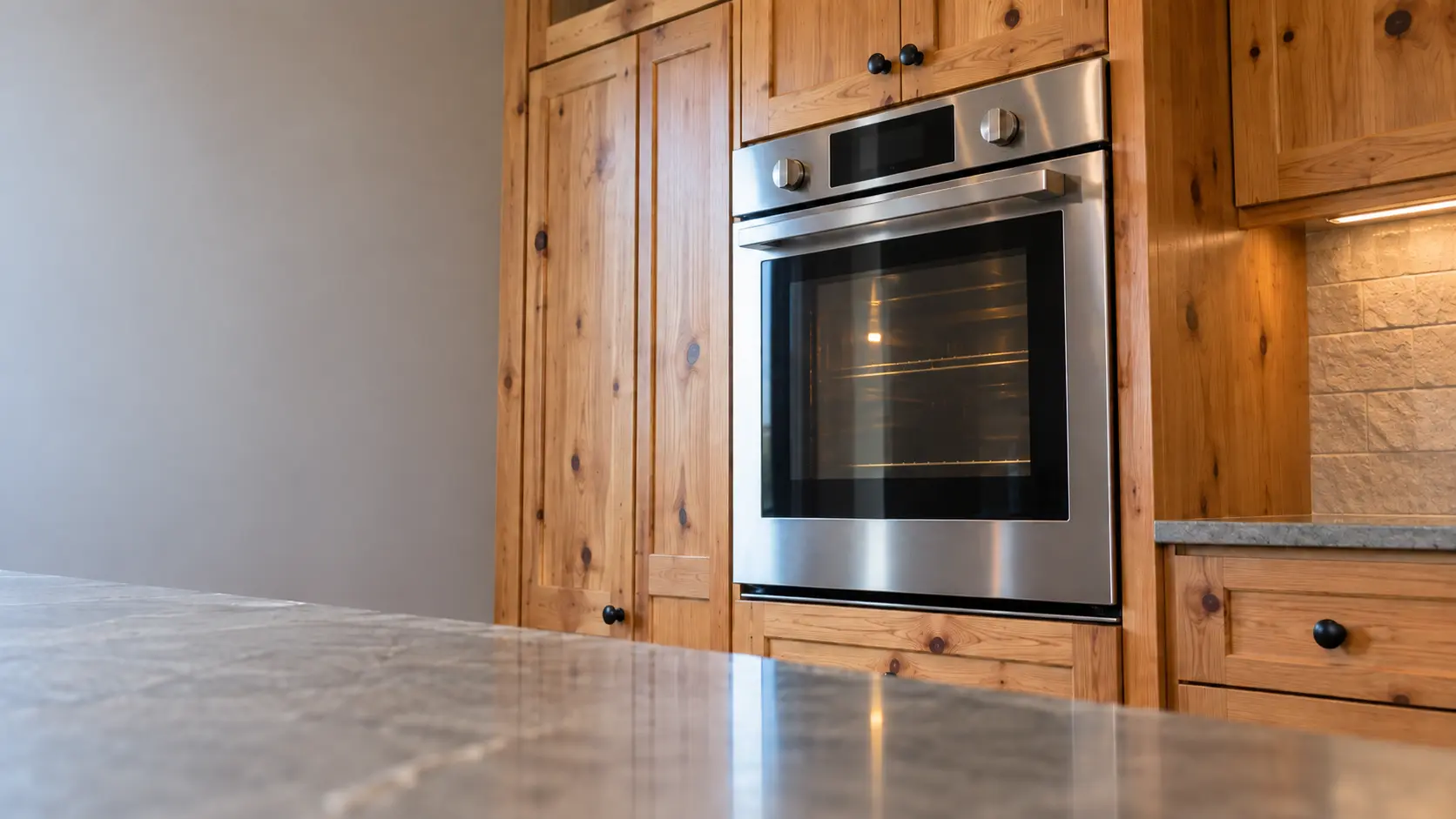 A kitchen wall with the oven in a rural ranch house kitchen with a wall oven in knotty pine cabinets, seen from a low angle, the oven door glass catching the light.