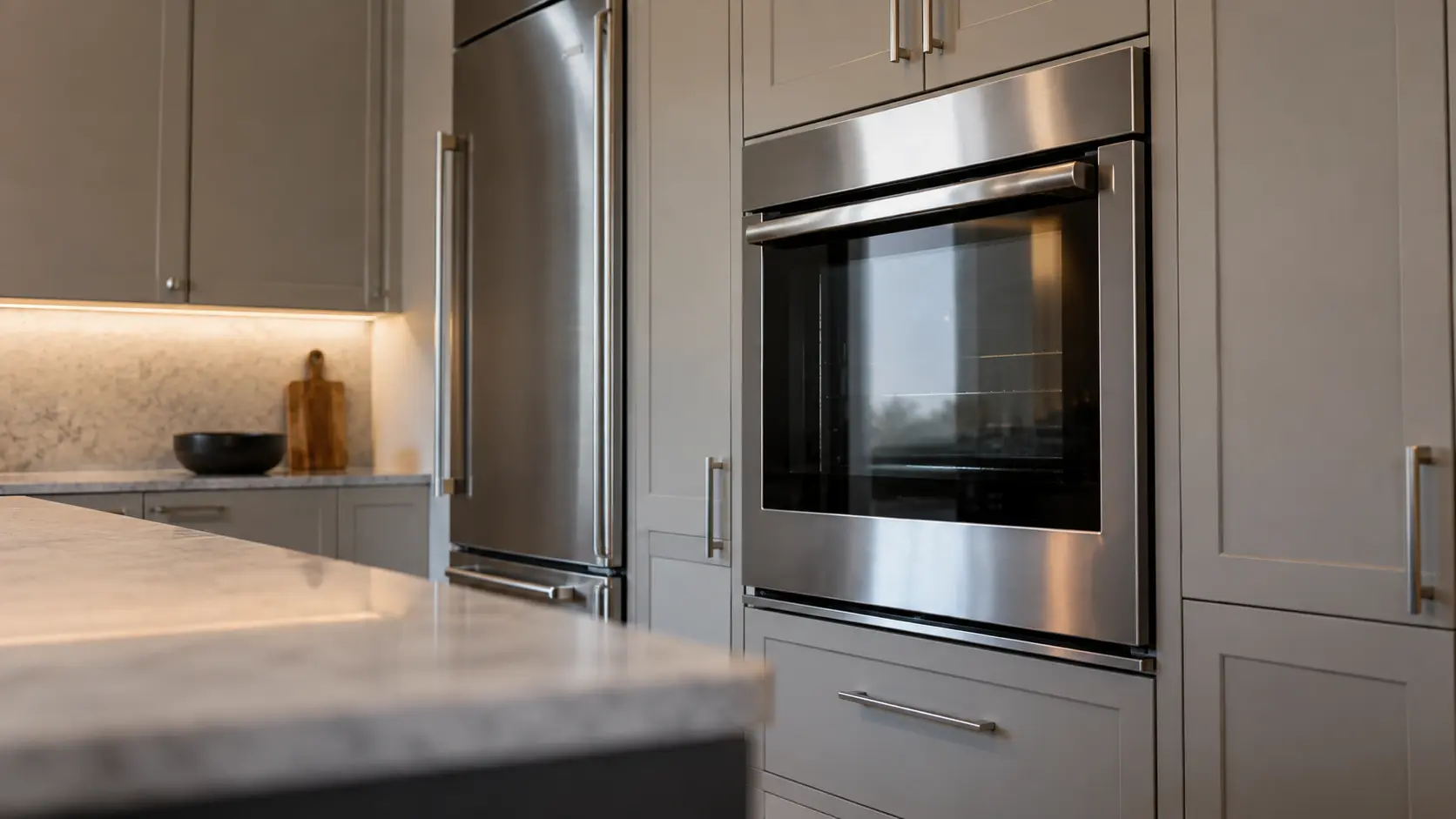 A kitchen wall with the oven in a townhouse kitchen with a wall oven beside the fridge column, seen from a low angle, the oven door glass catching the light.