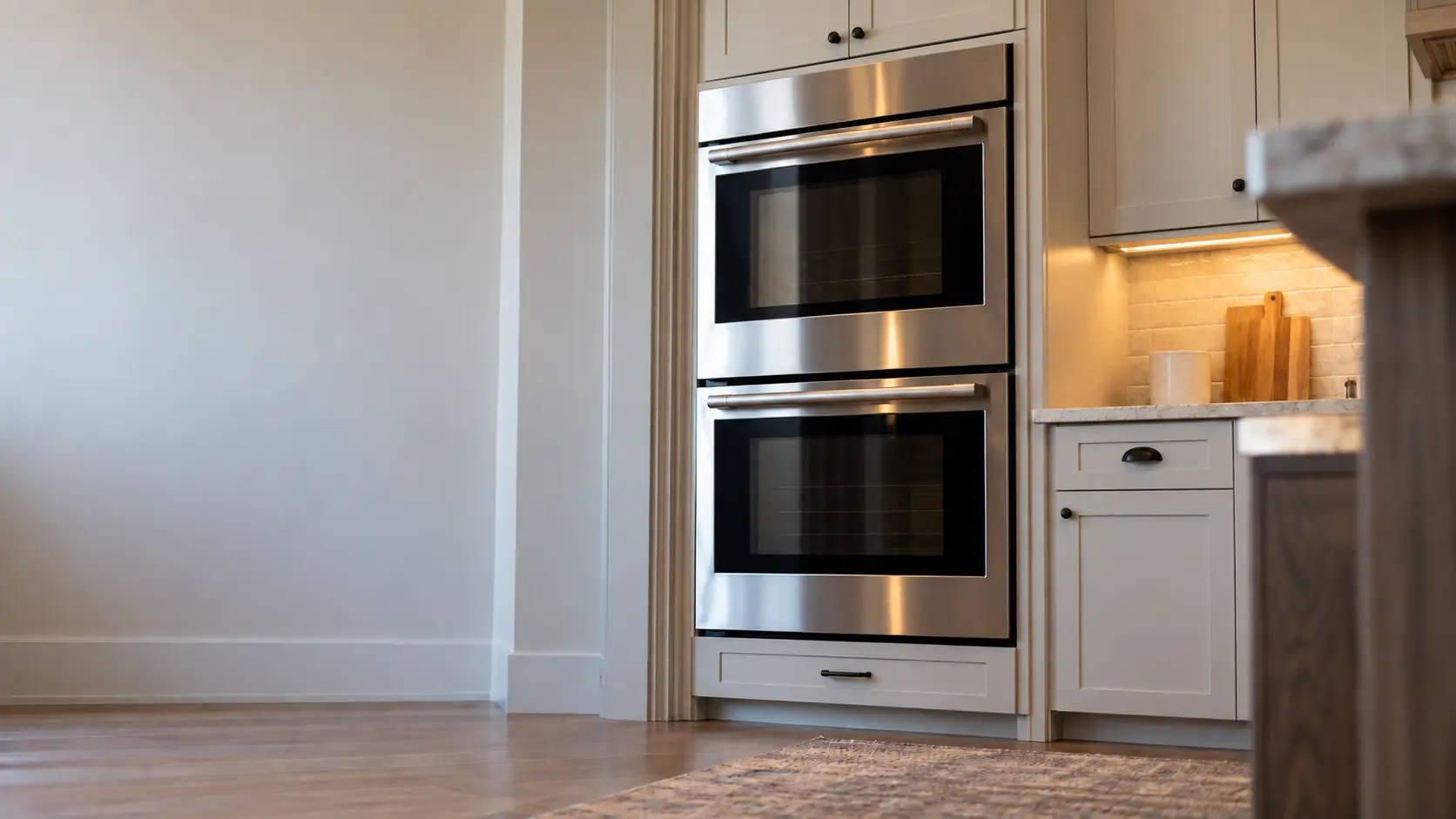 A kitchen wall with the oven in a vacation-rental kitchen with a double wall oven, seen from a low angle, the oven door glass catching the light.