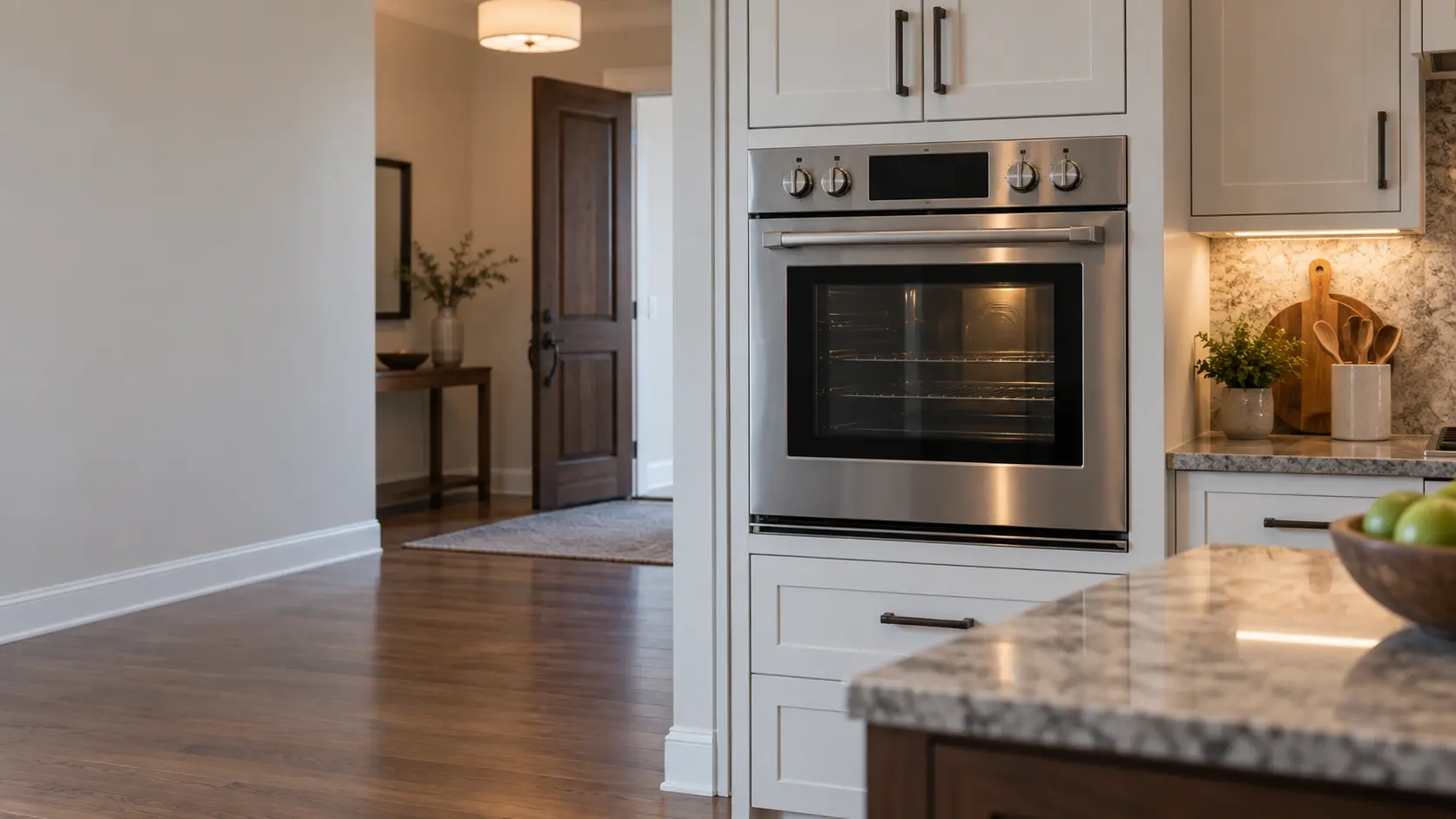 A US home kitchen with a stainless gas wall oven set into tall cabinets, the room empty and the front door open in the background as if the family has just stepped outside.