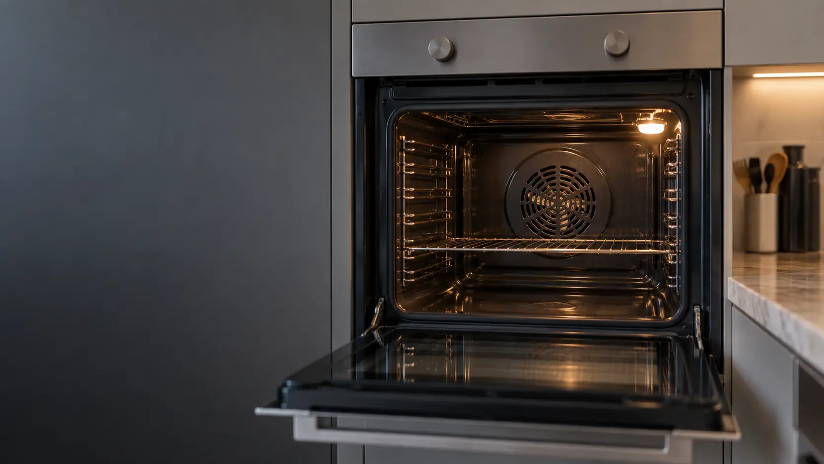 The round fan cover on the back wall of a convection wall oven, the blade of the fan faintly visible through its slots.