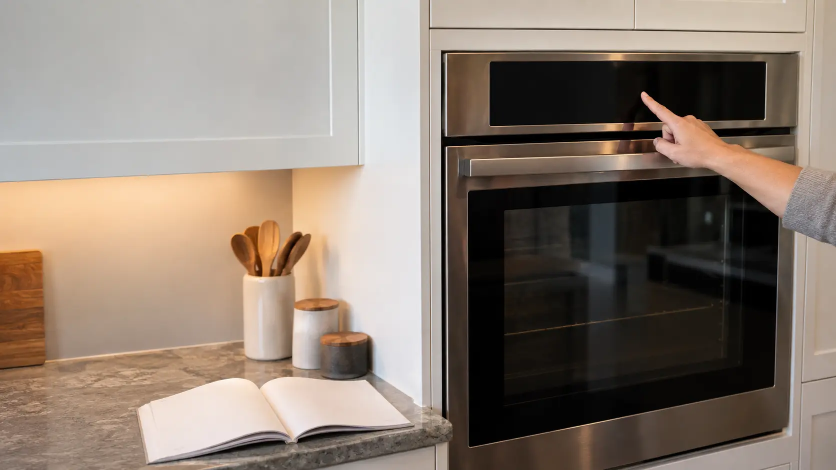 A built-in wall oven in a US home kitchen with its door closed and its display dark, a homeowner's finger pressing the touch controls above the door and the owner's manual open on the counter below.