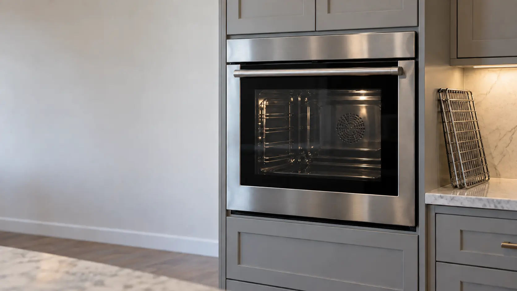 A stainless wall oven built into kitchen cabinetry in a US home, its door closed and latched with the window showing a clean empty cavity after a cleaning cycle, the oven racks leaning on the counter beside it.