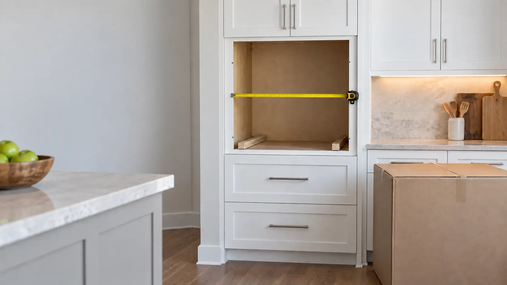 An empty wall oven opening in white shaker cabinetry in a US home kitchen, a tape measure stretched across the cutout and two level wooden runners at its base, with the new wall oven still boxed on the floor.